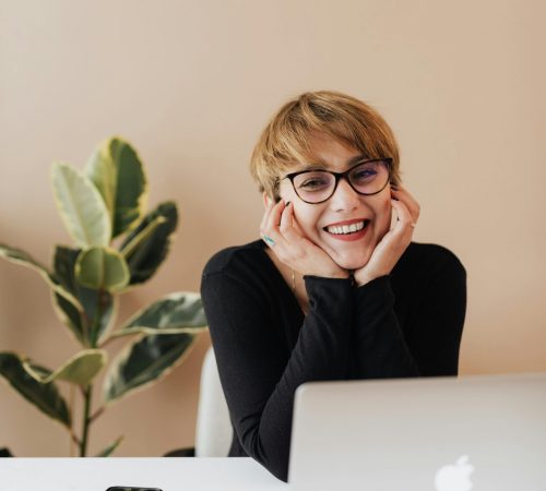 Cheerful woman in eyeglasses and black sweater smiling while working at a desk with a laptop indoors.